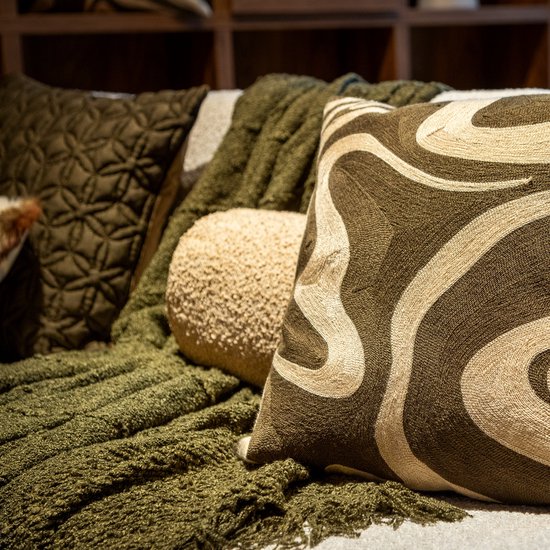 Detail van een olijfgroene plaid met franjes, naast een rond kussen en een kussen met marmerprint.