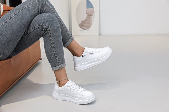 Een persoon in spijkerbroek, draagt witte lage sneakers met veters en een enkelbandje.