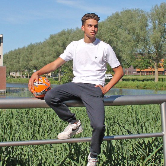 Jonge man zit op een reling met een voetbal, draagt een wit t-shirt en een donkerblauwe joggingbroek.