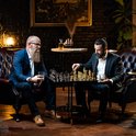 Twee mannen met baard spelen schaak in een luxe kamer. Één man houdt een glas whiskey vast.