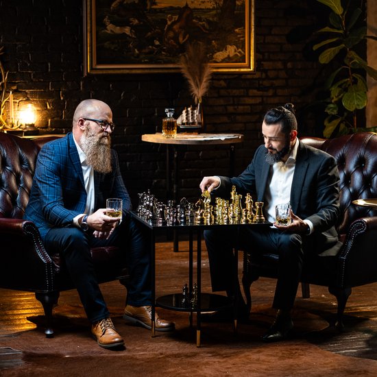 Twee mannen met baard spelen schaak in een luxe kamer. Één man houdt een glas whiskey vast.
