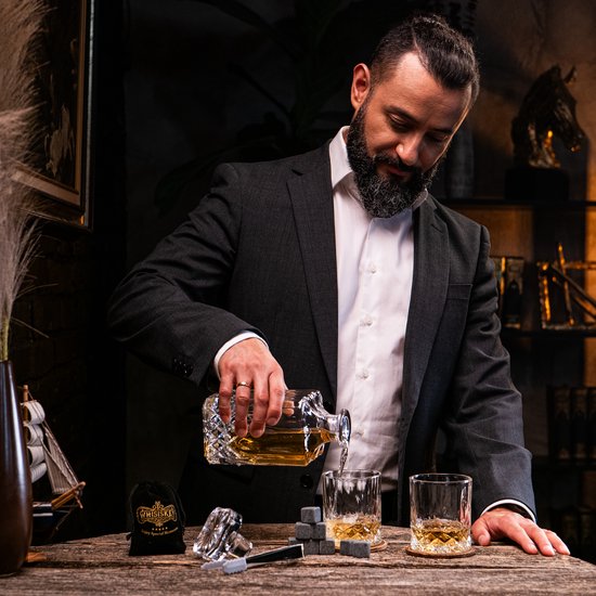 Man in pak schenkt whiskey uit een karaf in een glas. Whiskey stenen en zakje op de tafel.