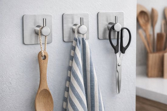 Drie zilverkleurige haken aan een witte muur, met een houten lepel, blauw gestreepte handdoek en schaar eraan.