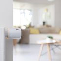 Netatmo slimme thermostaatknop, wit met oranje accent, op een witte radiator. Woonkamer op achtergrond.