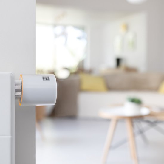 Netatmo slimme thermostaatknop, wit met oranje accent, op een witte radiator. Woonkamer op achtergrond.