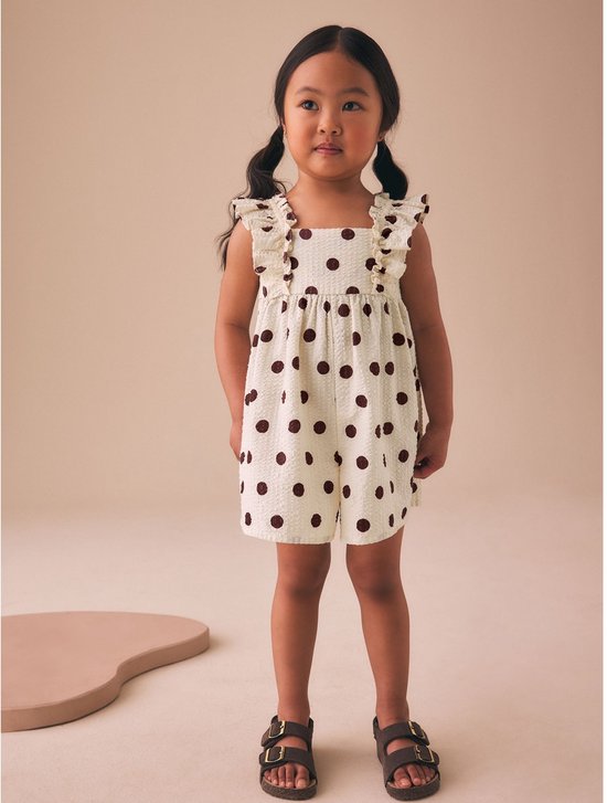 Meisje met staartjes staat in een crèmekleurige playsuit met bruine stippen, ruches en sandalen.