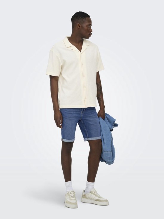 Man met korte mouwen, crèmekleurig hemd met open kraag. Blauwe jeans shorts, witte sokken en sneakers. Spijkerjasje in de hand.