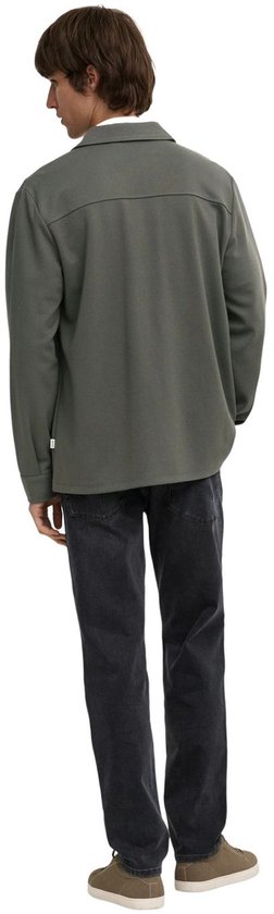 Man met de rug naar de camera, draagt olijfgroene utility blouse, grijze jeans en bruine sneakers.