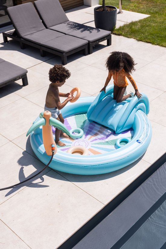 Kinderen spelen in een opblaasbaar zwembad met glijbaan. Een palmtak sproeier zorgt voor waterpret. Zonnige dag bij het zwembad.