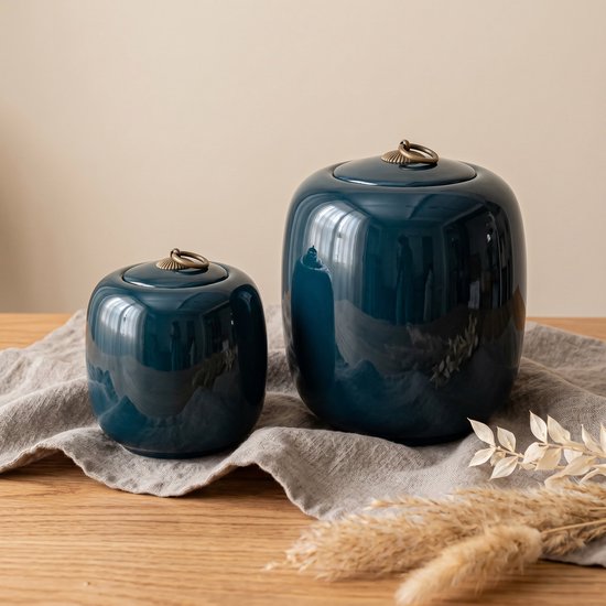 Twee donkerblauwe keramieken urnen met gouden ringen op de deksels, staand op een grijze doek.