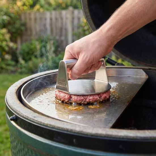 Hand gebruikt RVS smash burger pers om een burger te plat te drukken op een hete grill.