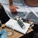 Groene fles zonnebrandcrème "The Green Junk SPF 30+" ligt op een open magazine op een houten tafel.
