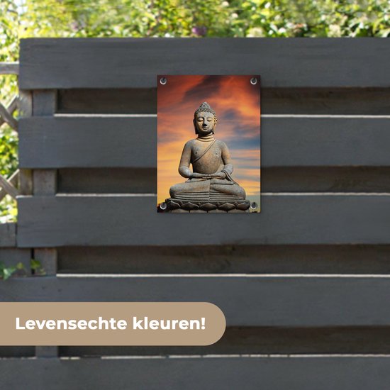 Bouddha en pierre sur fleur de lotus sur fond de coucher de soleil orange, suspendu à une clôture grise.