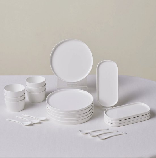 Service de table blanc composé d'assiettes empilées, de bols et de cuillères, avec un plat ovale sur le côté.