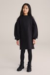 Een meisje met lang zwart haar draagt een zwarte oversized jurk met pofmouwen en een zwart logo op de borst, met een zwarte legging en zwarte laarzen.