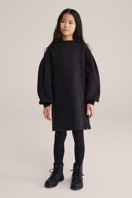 Een meisje met lang zwart haar draagt een zwarte oversized jurk met pofmouwen en een zwart logo op de borst, met een zwarte legging en zwarte laarzen.