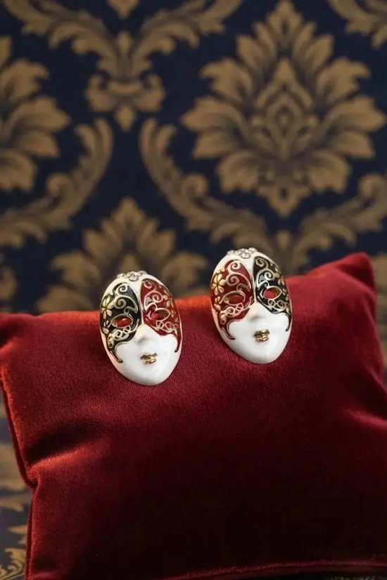 Paar oorbellen als witte Venetiaanse maskers met zwart-rode patronen en strass, gepresenteerd op een rood kussen.