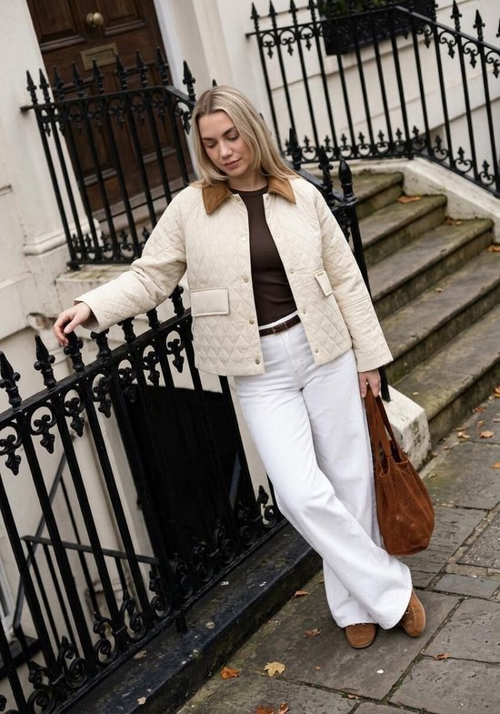 Vrouw in een gewatteerde crèmekleurige jas en witte wijde jeans, staand buiten.