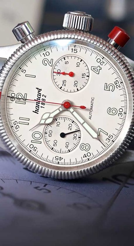 Close-up van een Hanhart Pioneer Mk II horloge met witte wijzerplaat en zwarte band. Zilverkleurige kast met chronograaffunctie en rode drukknoppen.