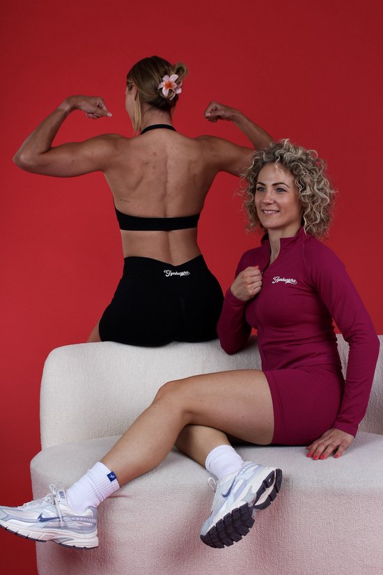 Twee vrouwen op een bankje voor rode achtergrond. Eén toont rugspieren in zwarte sportkleding, de ander zit in een bordeauxrode sportjurk.
