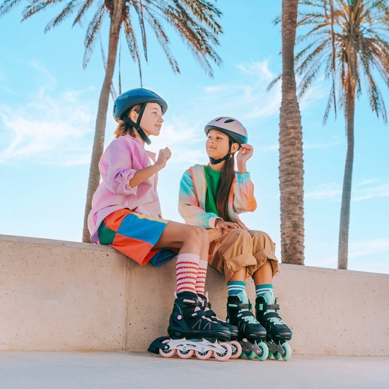 Twee kinderen met skeelers en helmen, zittend op een muur bij het strand.