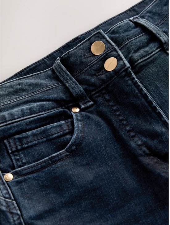 Detail van de hoge taille van een donkerblauwe spijkerbroek. De broek heeft een dubbele knoopsluiting met gouden knopen en een coin pocket.