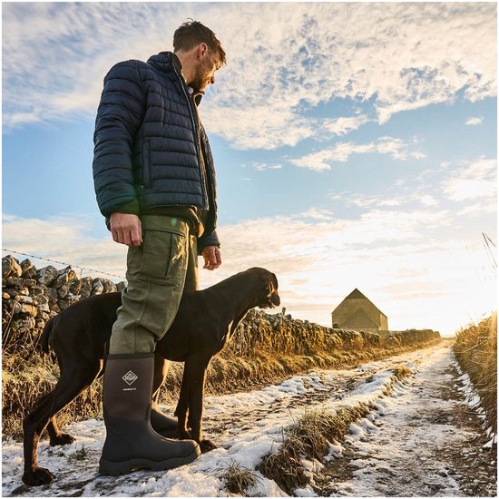 Een man met een hond staat op een modderig pad in de winter. Hij draagt de Muck Boot Derwent II laarzen.