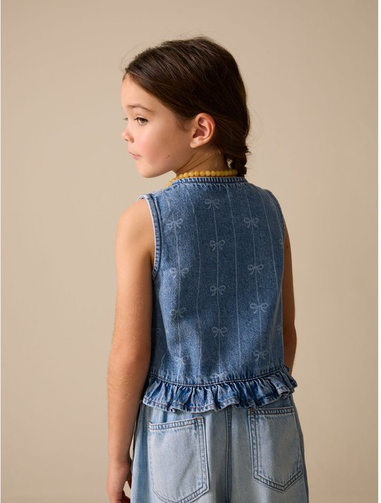 Meisje van achteren gezien, draagt een mouwloos denim gilet met strikprint en een geel kralensnoer.