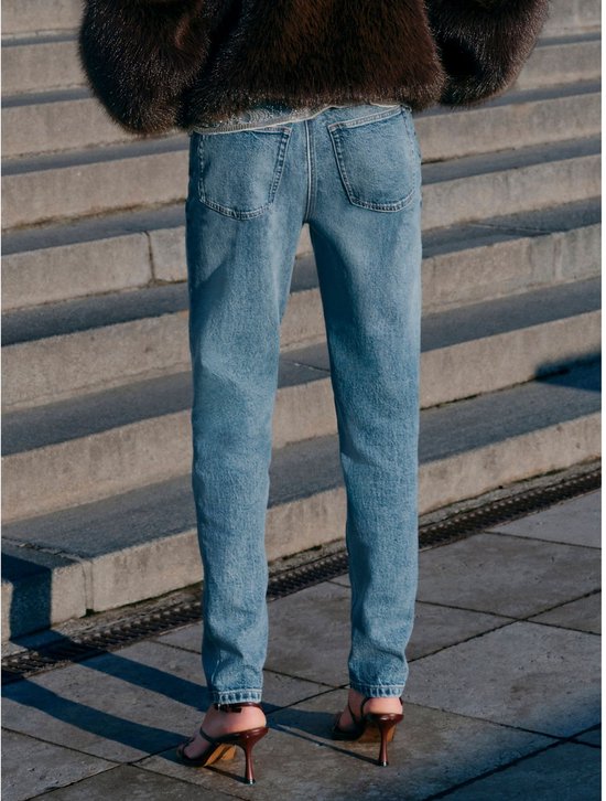 Model staand op een trap, draagt een spijkerbroek met hoge taille en hoge hakken, met een donkerbruine bontjas.