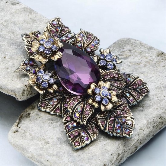 Gouden broche met grote paarse druppelvormige steen, omringd door kleine blauwe bloemen en paarse strass steentjes.