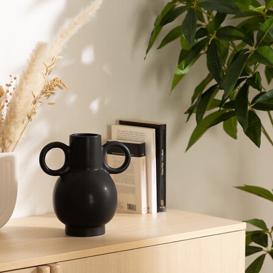 Zwarte vaas met ronde handvatten op een houten kastje naast boeken en gedroogde pampasgrassen.