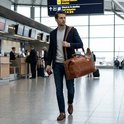Man met blauwe blazer, trui en bruine tas loopt door een luchthaven met Gate-bord.