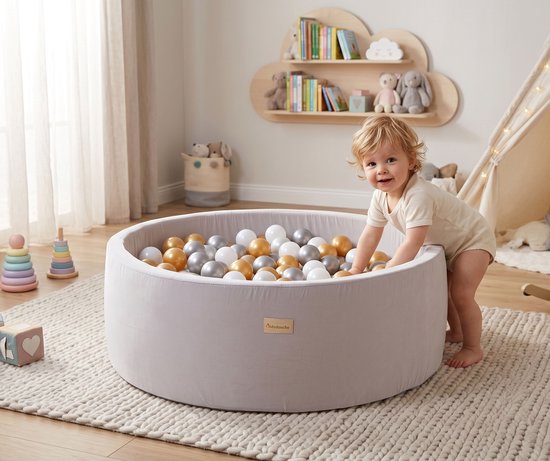 Een peuter speelt in een taupe grijze ballenbak met gouden, zilveren en parelmoer ballen. De peuter staat op een beige geruit kleed.
