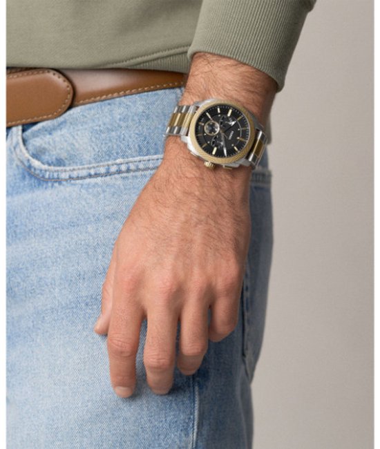 Gouden en zilverkleurig Fossil horloge om een mannelijke pols, gedragen met spijkerbroek en bruine riem.