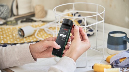 Handen gebruiken de Bosch Zamo afstandsmeter met lintadapter om de omtrek van een metalen rek te meten.
