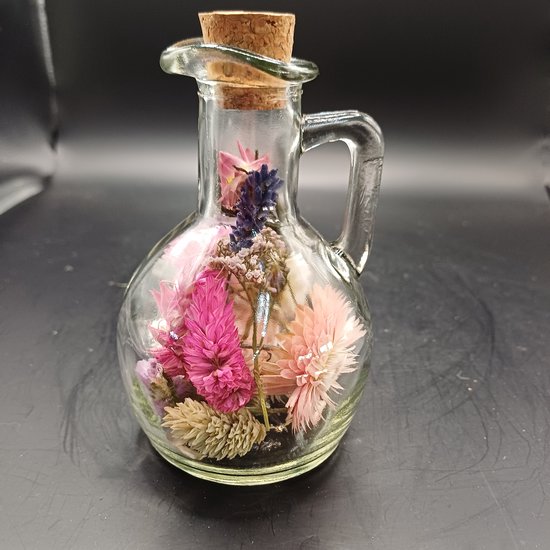 Glazen karafje met handvat, gevuld met gedroogde bloemen in roze, paarse en witte tinten, afgesloten met een kurk.