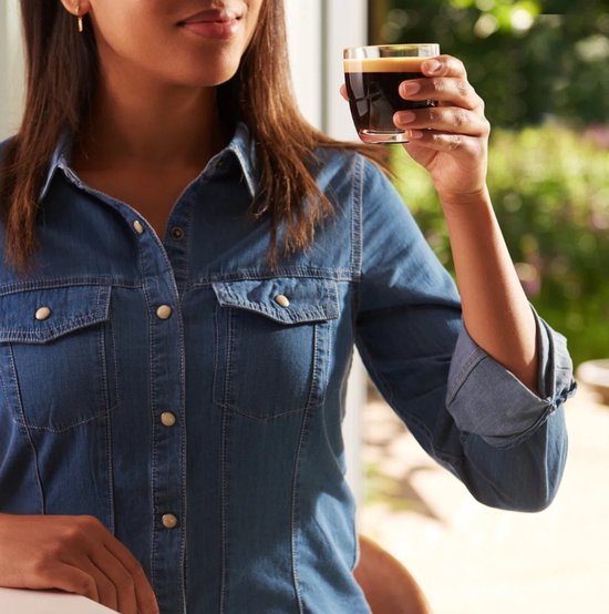Een vrouw in spijkerblouse houdt een glas espresso omhoog.