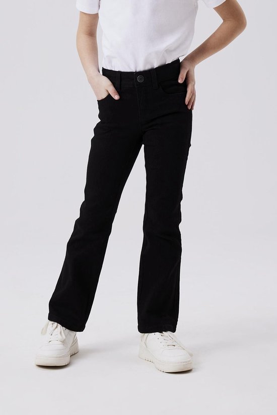 Meisje draagt een zwarte flared jeans en witte sneakers, met handen in de zakken.