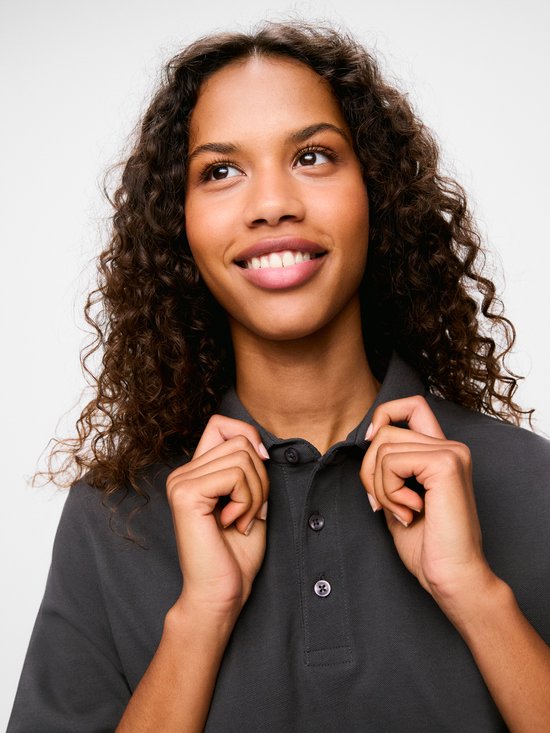 Vrouw met krullend haar, draagt een donkergrijs poloshirt en trekt aan de kraag.