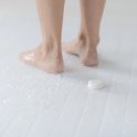 Voeten op een witte douchetegelvloer, met waterdruppels en een douchetablet op de vloer.