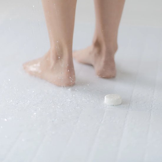 Voeten op een witte douchetegelvloer, met waterdruppels en een douchetablet op de vloer.