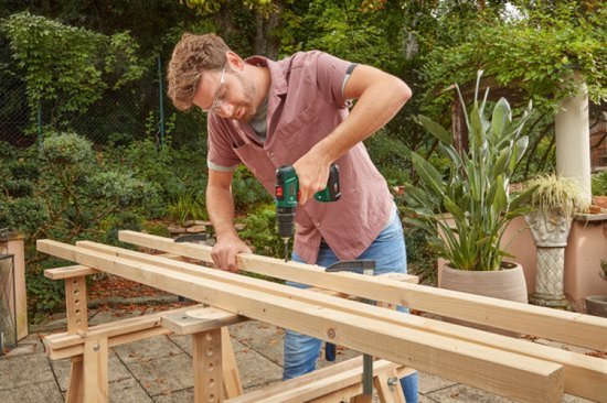 Man met bril en roze hemd boort in houten balken met een groene Bosch accuboorschroevendraaier.