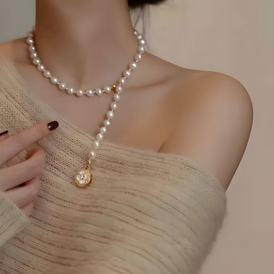 Crèmekleurige parelketting met een gouden bloemvormige hanger met zirkoon en parelmoer, gedragen door een vrouw met een beige trui.