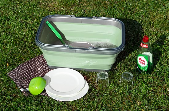 Groene opvouwbare wasteil met een groene borstel erin, gevuld met water en zeep. Een afwasmiddelfles en borden staan ernaast op het gras.