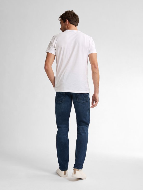 Man met de handen in de zakken, staand in een donkerblauwe, slimfit jeans en een witte T-shirt, van achteren gezien.