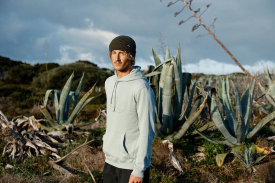 Man met Nomad-muts in mintgroene hoodie staat voor planten in een natuurlijke omgeving.