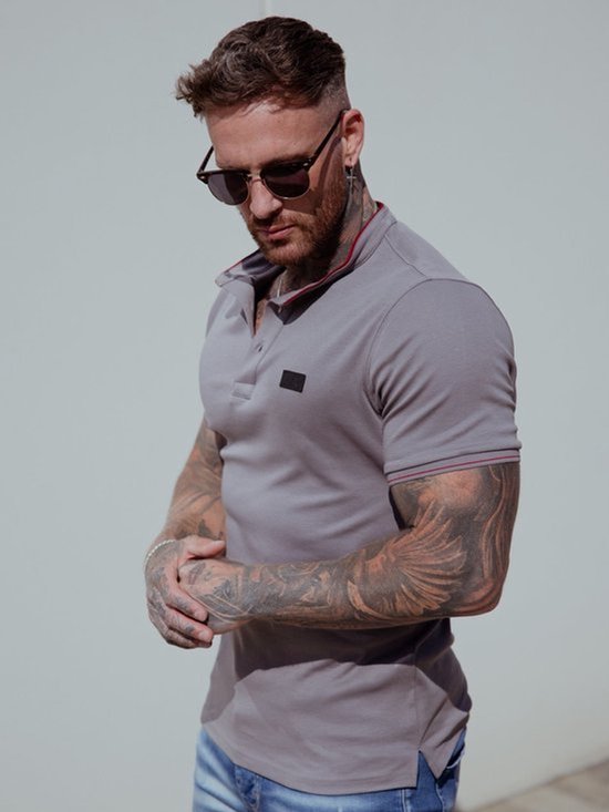 Man met tatoeages draagt een lichtgrijze polo met rode bies langs de kraag en mouwen. Hij draagt een zonnebril.