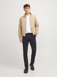 Man staat, draagt een beige bomberjack en een donkerblauwe chinobroek.
