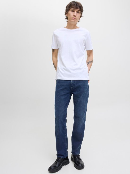 Jonge man met tatoeages in een witte T-shirt en donkere jeans met handen in de zakken.