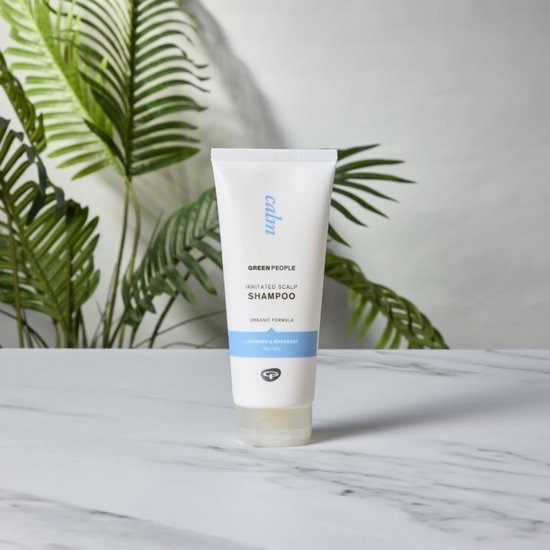 Witte tube Green People Irritated Scalp Shampoo, met blauwe band en 'lavender & rosemary', op een marmeren ondergrond met planten op de achtergrond.